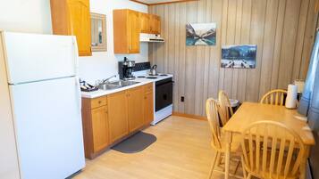 A compact kitchen with wooden cabinets, a white refrigerator, a stove, and a sink. There are two wooden chairs around a small table.