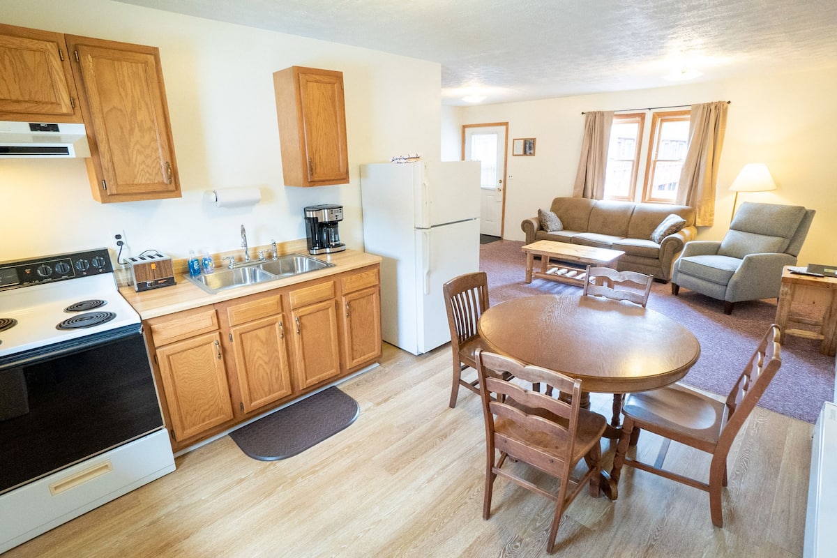 A compact kitchen with wooden cabinets, a stove, and a refrigerator. There is a dining area with a table and chairs, and a living room with a sofa and a lamp.