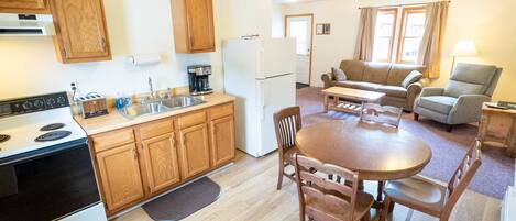 Cocina compacta con armarios de madera, estufa y refrigerador. Hay un comedor con mesa y sillas, y una sala de estar con sofá y lámpara.