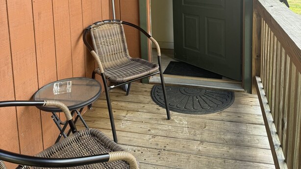 Eine hölzerne Veranda mit zwei Korbstühlen, einem kleinen Tisch, einer Tür mit einem Kranz und einem Holzgeländer.