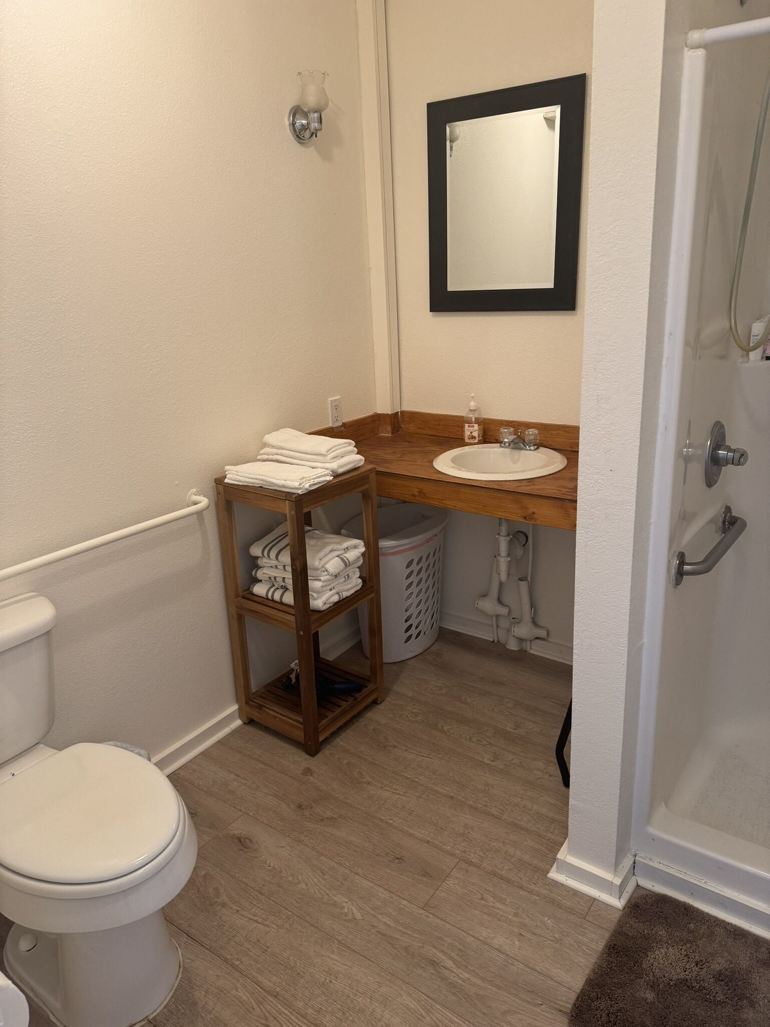 Un baño con inodoro, lavamanos y ducha. Hay un perchero con toallas dobladas y un cesto de ropa.