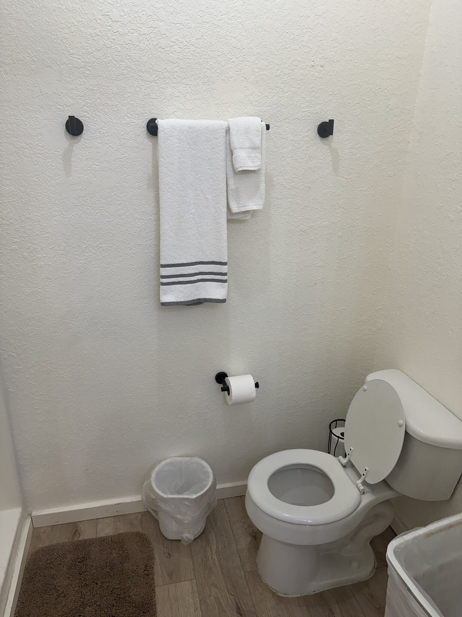 Een badkamer met toilet, prullenbak, handdoekrek met twee handdoeken en toiletpapierhouder.