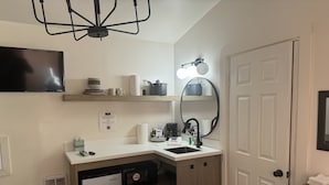 Una cocina compacta con lavavajillas, un refrigerador pequeño, un lavabo y una encimera con diversos objetos. Se aprecia un accesorio de iluminación en el techo y un espejo adherido a la pared sobre el lavabo.