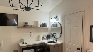 Une cuisine compacte équipée d’un lave-vaisselle, d’un petit réfrigérateur, d’un évier et d’un plan de travail sur lequel sont disposés divers objets. Un luminaire est fixé au plafond et un miroir est accroché au mur au-dessus de l’évier.