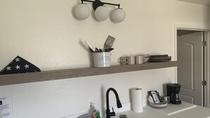 A kitchen with a sink, a coffee maker, and a shelf with mugs and utensils.