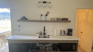 A kitchen with a white countertop, a sink, a microwave, a coffee maker, and a wooden shelf above.