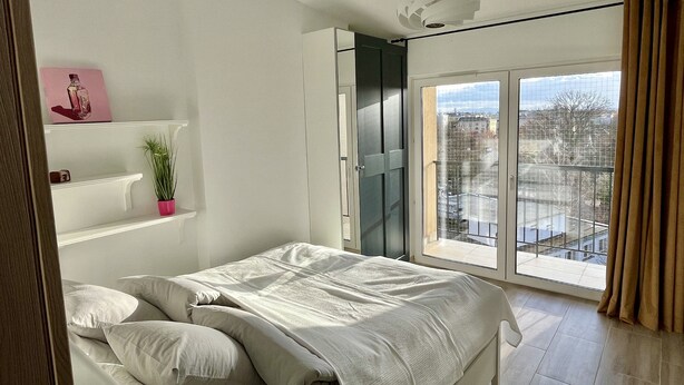 Un dormitorio con una cama, una planta en maceta, una estantería con un cuadro y vistas de la ciudad a través de un ventanal grande.