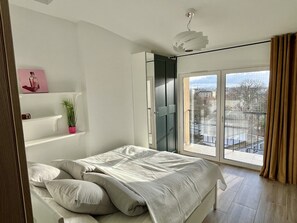 A bedroom with a bed, a potted plant, a shelf with a picture, and a view of the city through a large window. - Ferienwohnung Ruhig, Zentrumsnah (Kraków)