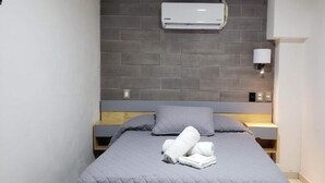 A small, neatly arranged bedroom with a grey bed, a grey pillow, and a neatly folded white towel on the bed. There is a wall-mounted air conditioner above the bed and a small bedside table with a lamp. - El Suburbio - Morelos Sur (Morelia)