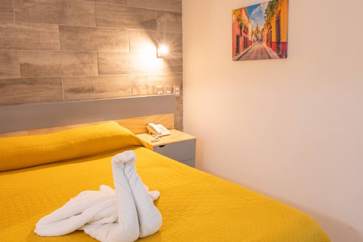 A hotel room with a large bed, a wooden headboard, a painting on the wall, and a bedside table with a lamp.