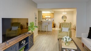 Ein Wohnzimmer mit Fernseher, einer Couch, einem Couchtisch mit Blumen und einer Küche im Hintergrund.