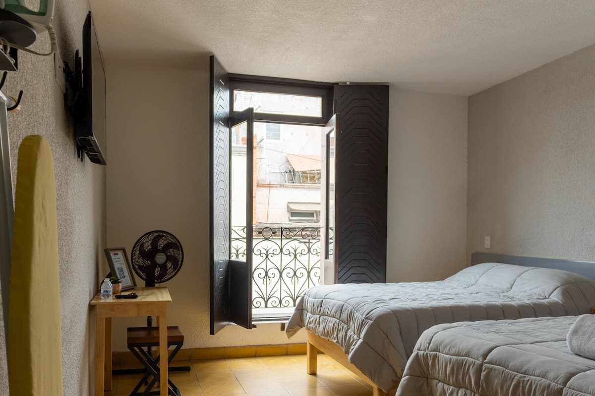 A bedroom with two beds, a wooden desk, a standing fan, and a window with a view of buildings.