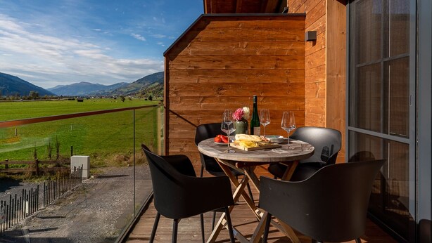 Un balcón de madera con una mesa dispuesta para dos personas, con vistas a un campo verde y montañas.