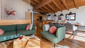 Una sala de estar moderna con sofás verdes, vigas de madera, una zona de comedor y una cocina al fondo.
