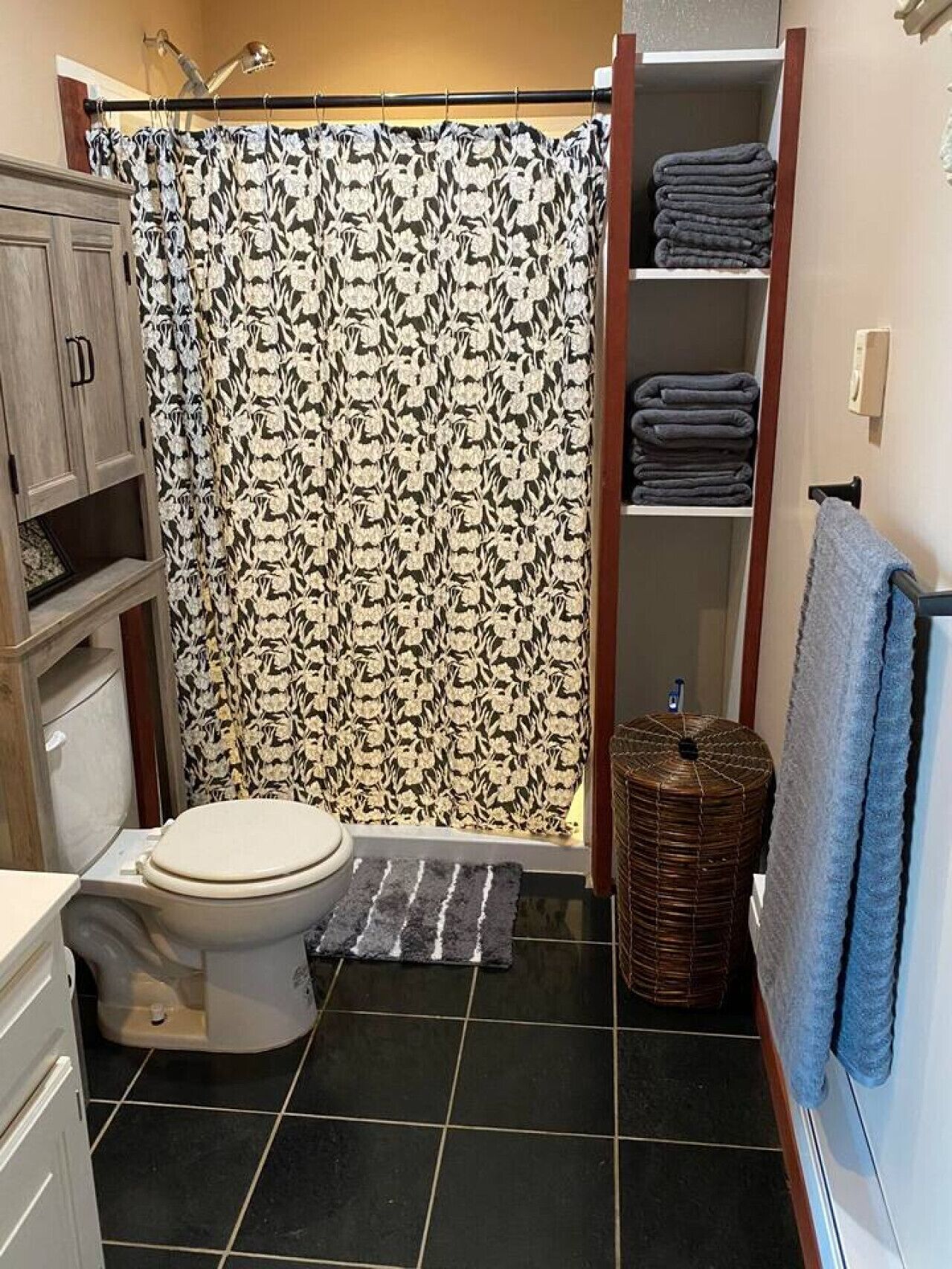 Une salle de bain comprenant des toilettes, un rideau de douche à motifs floraux, un porte-serviettes et une étagère avec des serviettes pliées.