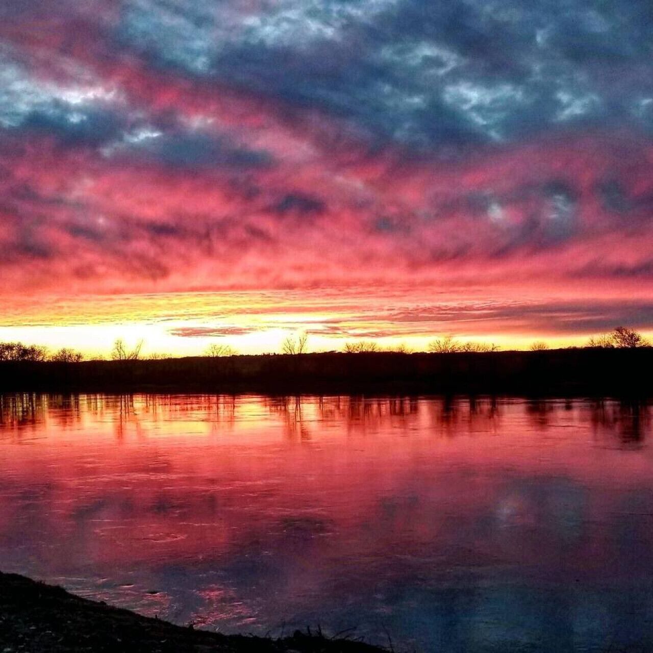 A sunset over a calm body of water with a silhouette of trees in the distance.