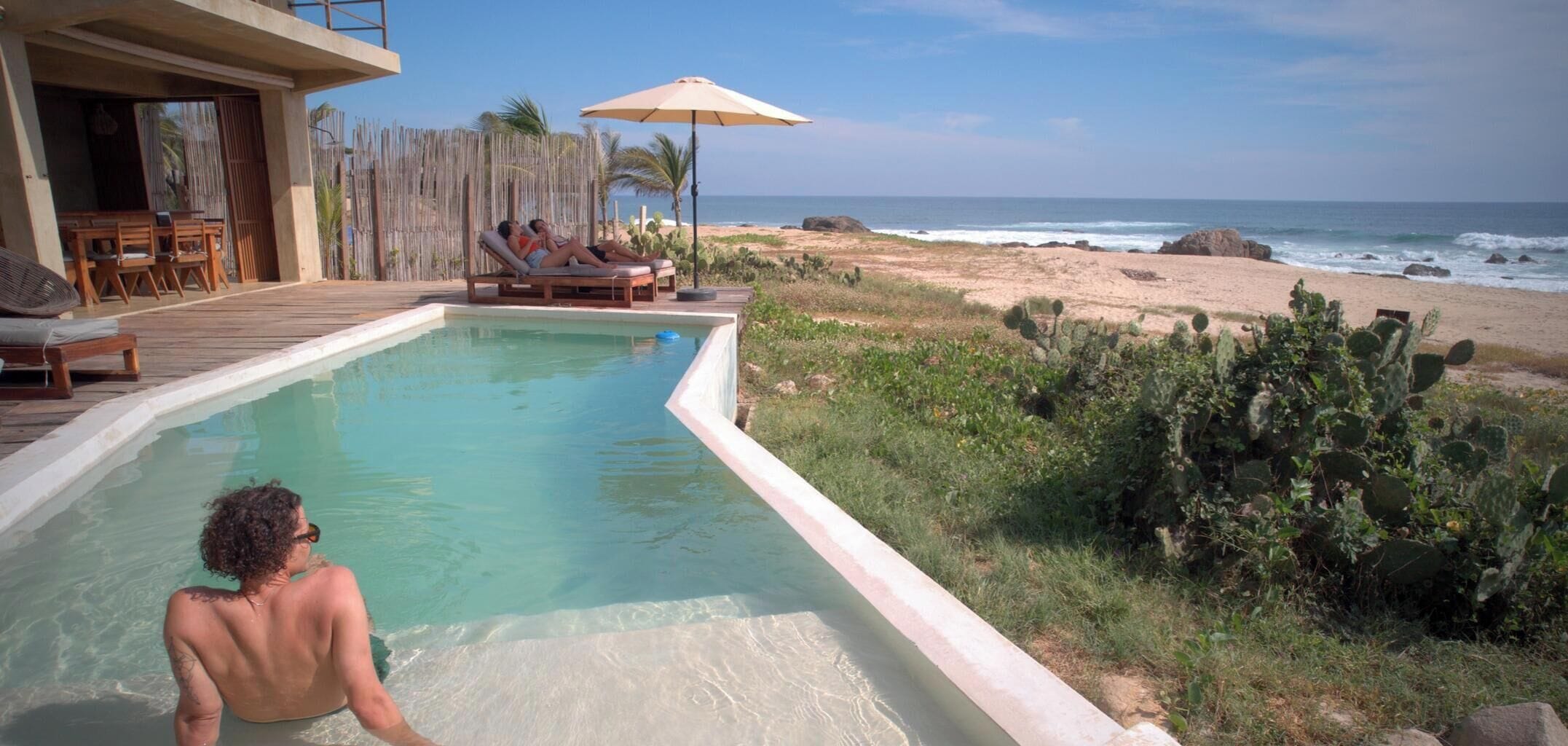 A pool with a view of the beach and ocean, wooden deck chairs, and a wooden fence.