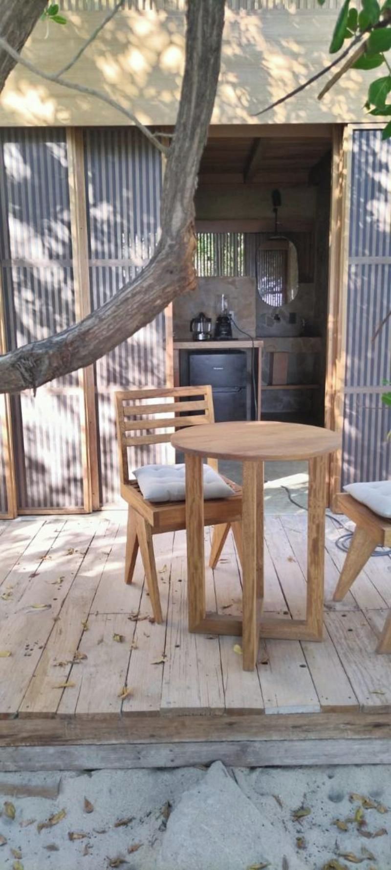 Eine Holzterrasse mit einem runden Tisch und zwei Stühlen, ein Baumzweig darüber und eine Küchenzeile im Hintergrund.