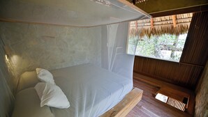 Un lit simple, protégé par une moustiquaire, dans une chambre rustique aux murs de pierre et au plancher en bois.