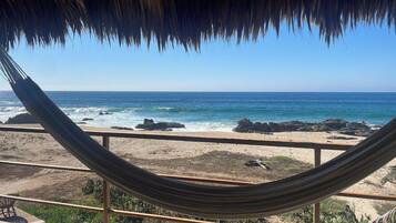 A hammock under a thatched roof overlooking a beach with rocky shoreline and calm sea.