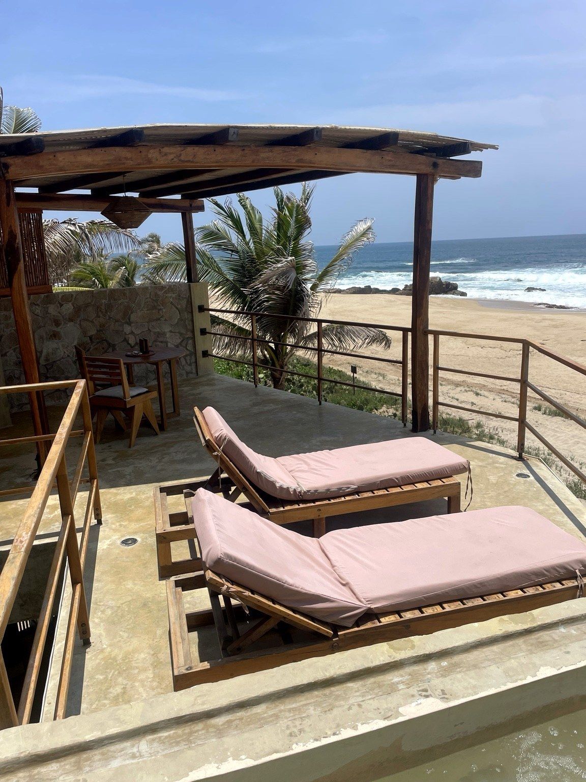 A beachside lounge with wooden loungers, a stone wall, and a view of the ocean.