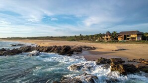 A beachfront property with a thatched-roof house, palm trees, and rocky shoreline.