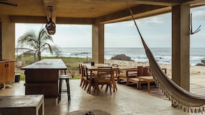 A beachside dining area with a wooden table and chairs, a hammock, and a view of the ocean.
