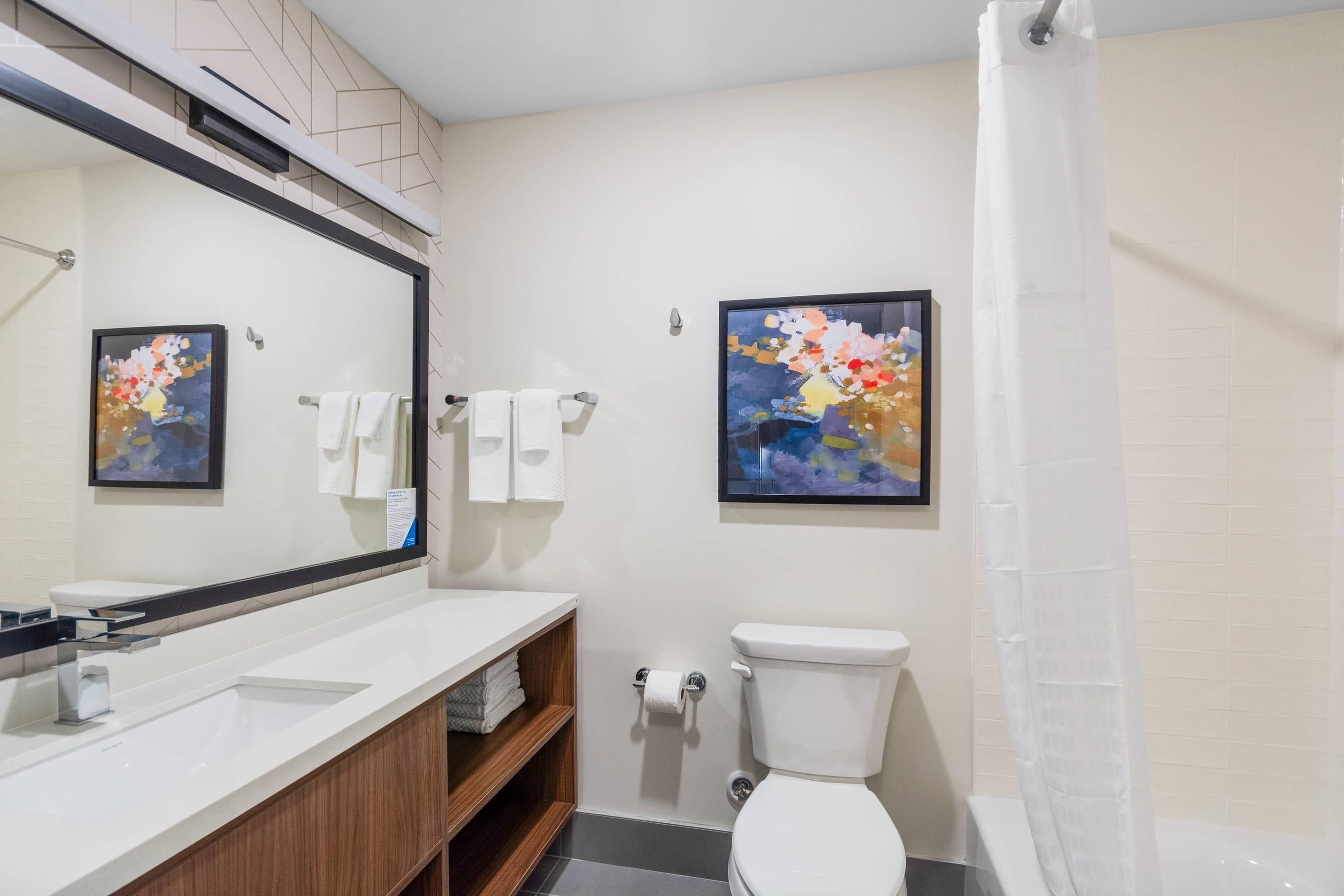 Un baño moderno con lavamanos blanco, inodoro y cortina de ducha. Hay dos cuadros enmarcados en la pared.