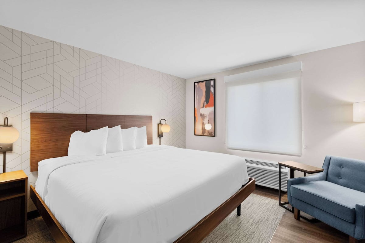 Una habitación de hotel con una cama grande, un sillón azul, una mesita de noche con lámpara y una obra de arte colgada en la pared.
