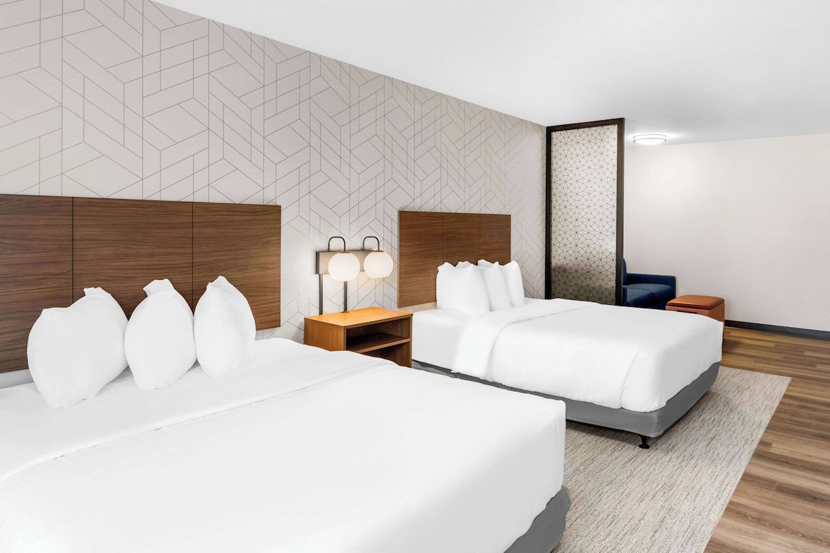Una habitación de hotel moderna con dos camas, un cabecero de madera, una mesita de noche con lámpara y un panel decorativo en la pared.