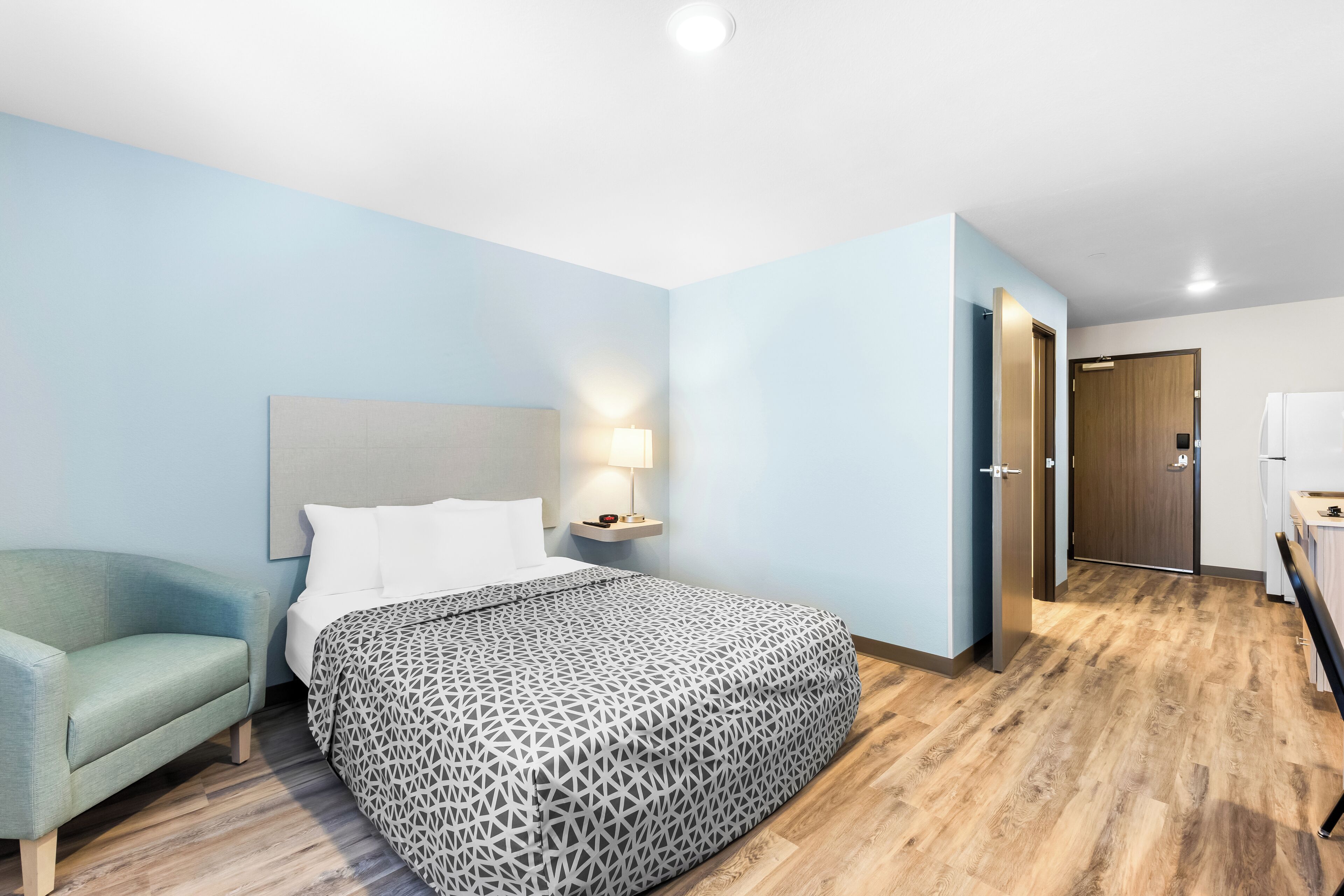 A hotel room with a bed, a nightstand, a lamp, a chair, and a wooden floor.