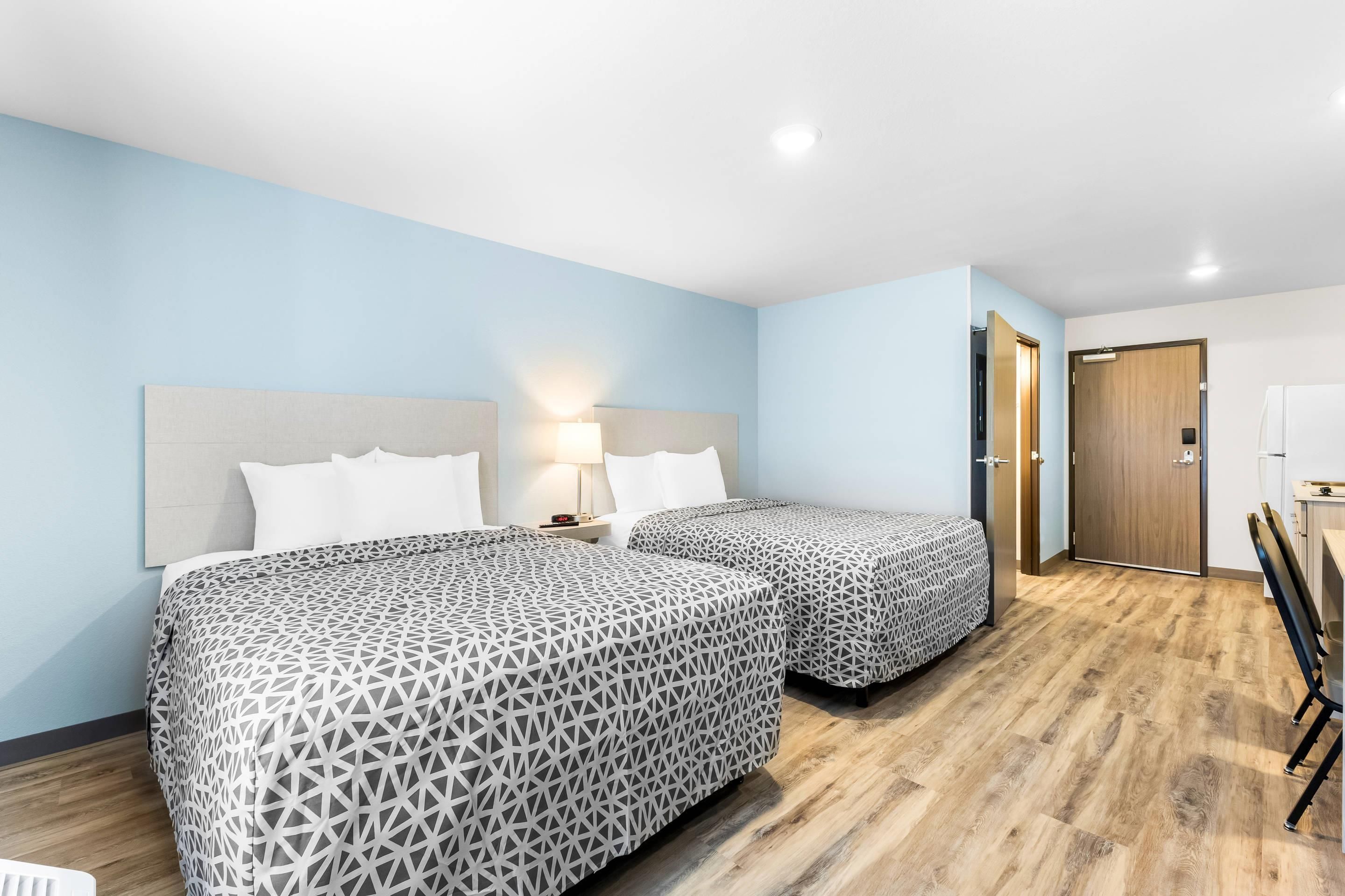 A hotel room with two beds, a nightstand, a lamp, a wooden floor, and a door leading to a bathroom.