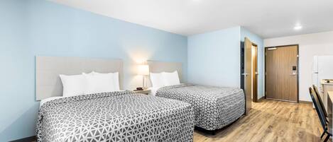A hotel room with two beds, a nightstand, a lamp, a wooden floor, and a door leading to a bathroom.
