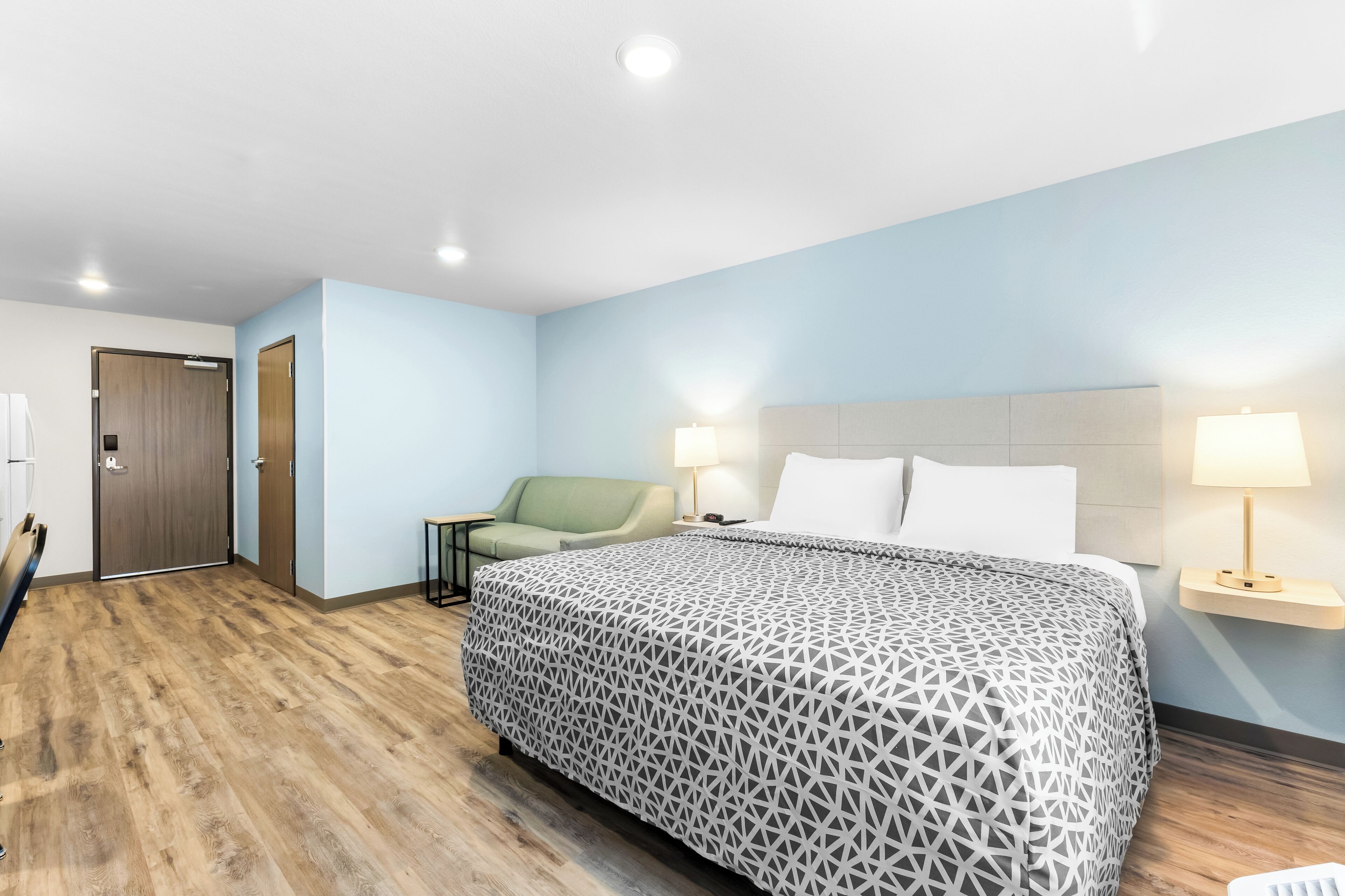 Une chambre d’hôtel avec un grand lit, un fauteuil vert, un plancher en bois et une porte.