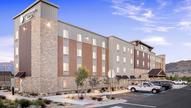 A hotel with a stone facade, multiple windows, and a parking lot with several vehicles. - Woodspring Suites Bluffdale Salt Lake City (Bluffdale)