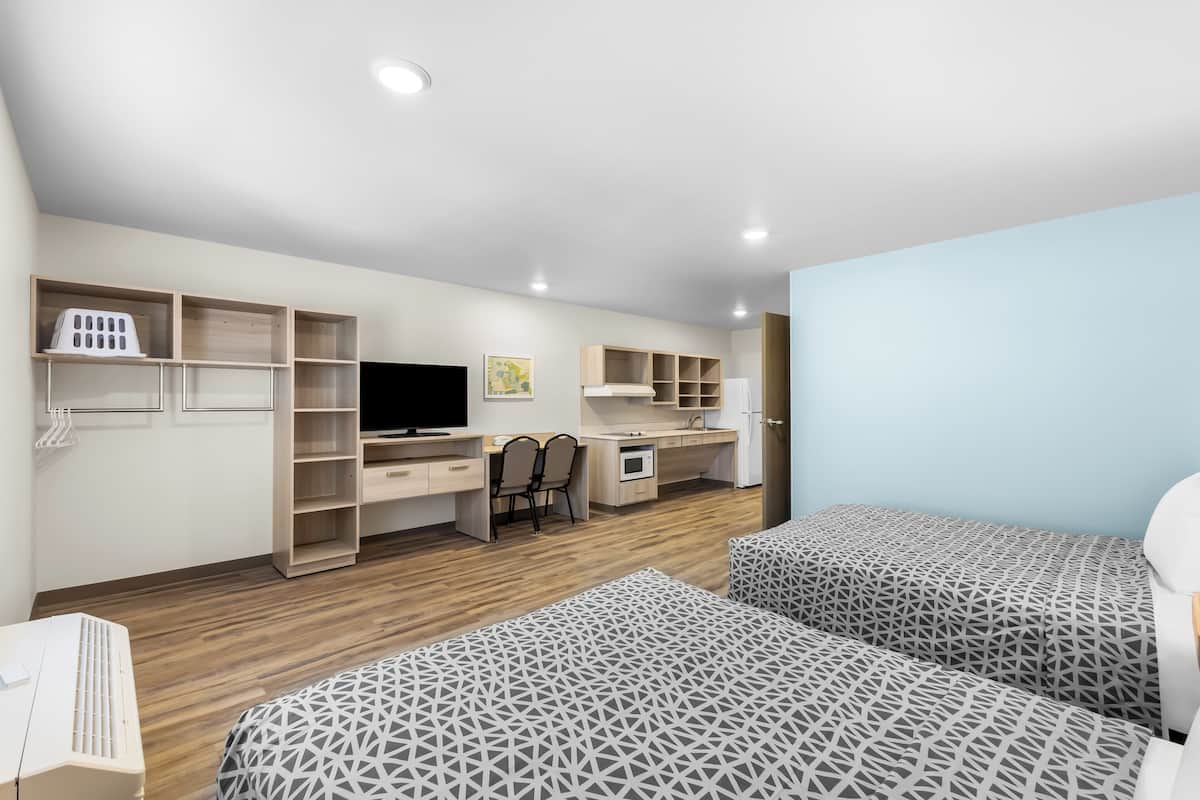 A modern hotel room with two beds, a desk, a TV, and built-in shelving.