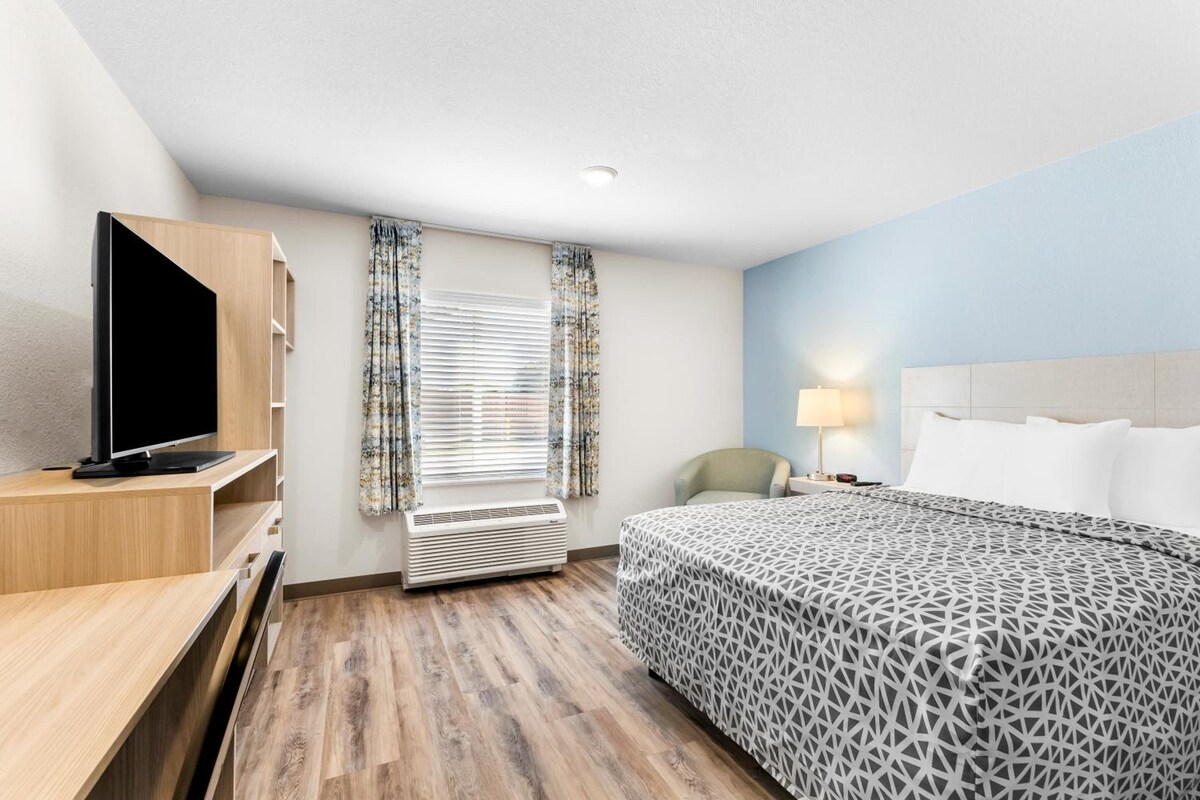 A hotel room with a bed, a desk with a television, a chair, a window with curtains, and a wall-mounted air conditioner.
