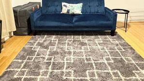 A dark blue sofa with a patterned throw pillow, a grey and white rug, and a small black side table with a plant. - ⭐Cozy 1-Bed⭐Minutes to NYC/PATH (Jersey City)