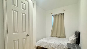A bedroom with a bed, a window with a curtain, a door, and a trash bin. - ⭐Cozy 1-Bed⭐Minutes to NYC/PATH (Jersey City)