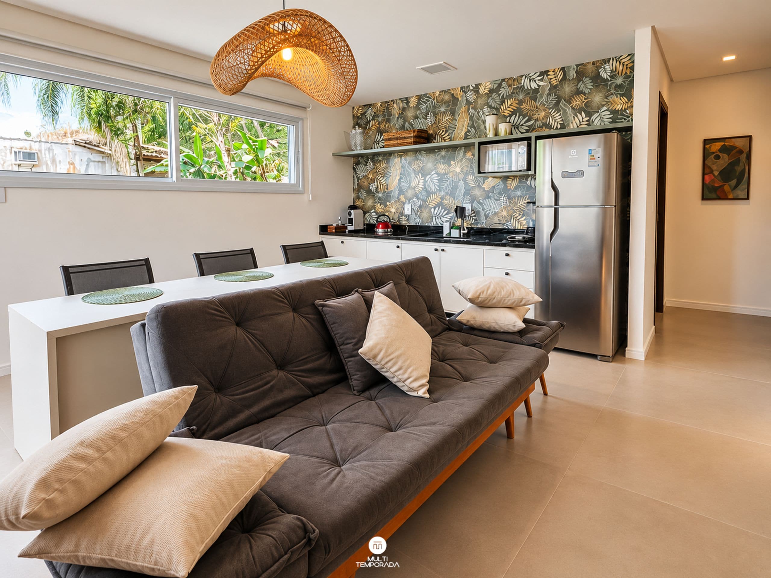 A modern kitchen with a built-in refrigerator, a dining area with a sofa and chairs, and a large window with a view of greenery.
