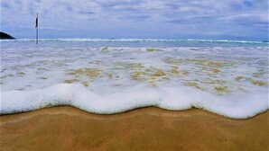 A beach with waves gently lapping at the shore, a flagpole in the distance, and a cloudy sky. - Villa Verde Bangalô - Garopaba - SC (Garopaba)