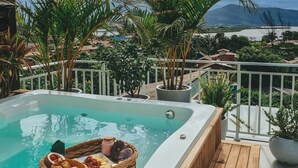 A hot tub with a wicker basket filled with snacks, surrounded by potted plants and overlooking a scenic view of a lake and mountains. - Villa Verde Bangalô - Garopaba - SC (Garopaba)