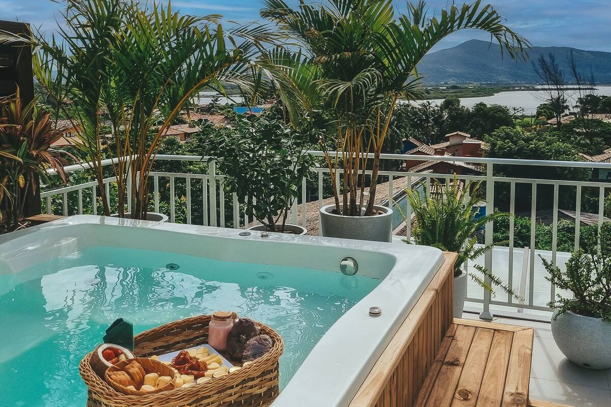 A hot tub with a wicker basket filled with snacks, surrounded by potted plants and overlooking a scenic view of a lake and mountains.