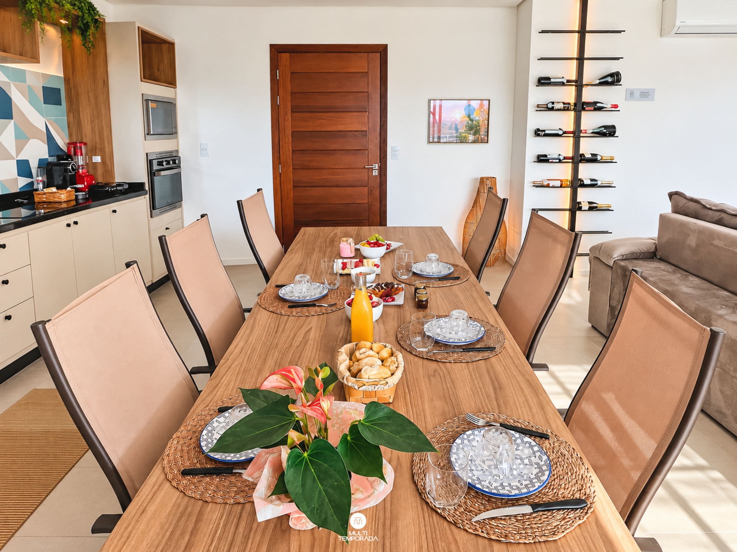 A dining area with a wooden table set for six, surrounded by chairs, with a kitchen and a wine rack visible in the background.