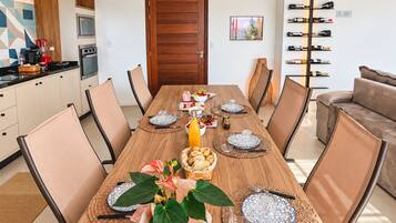 A dining area with a wooden table set for six, surrounded by chairs, with a kitchen and a wine rack visible in the background.