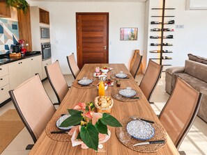 Un comedor con una mesa de madera dispuesta para seis personas, rodeada de sillas, con una cocina y una vinoteca visibles al fondo.