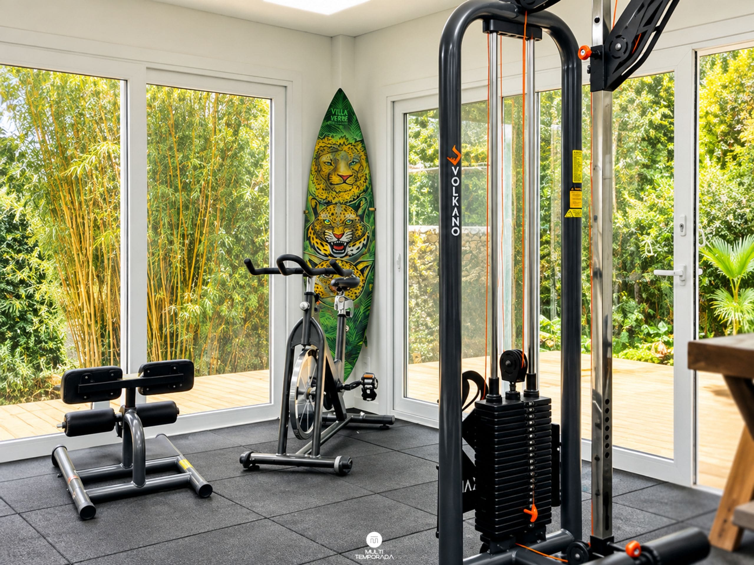 A gym with exercise equipment, a yoga mat, and a view of greenery through large windows.