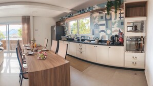A modern kitchen with a curved island, a dining area with a sofa, and a view of a balcony. - Villa Verde Bangalô - Garopaba - SC (Garopaba)