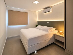 A hotel room with a bed, bedside table, lamp, air conditioning unit, and a window with blinds. - Villa Verde Bangalô - Garopaba - SC (Garopaba)
