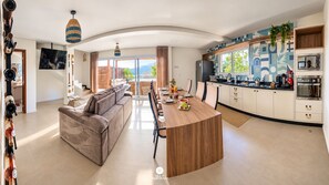 A modern kitchen with a central island, dining area with a sofa, and a large window offering a view. - Villa Verde Bangalô - Garopaba - SC (Garopaba)
